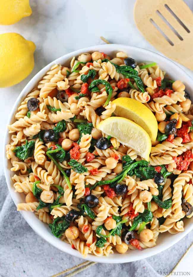 overhead shot of Vegan Pasta Salad in white bowl with grey napkin, lemons and wooden tongs