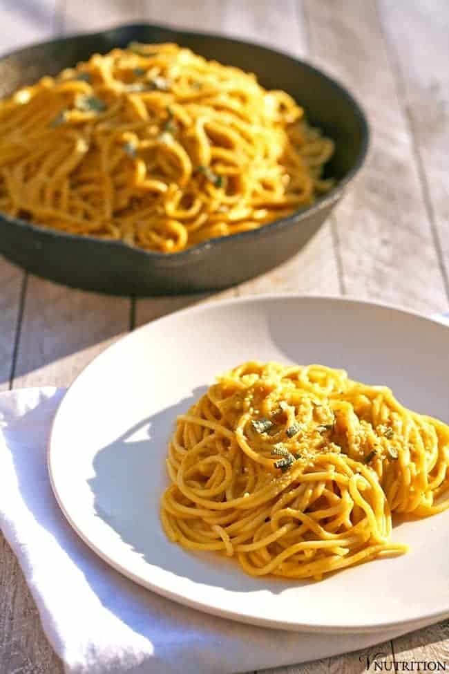 a serving of Vegan Pumpkin Pasta topped with vegan parmesan on a white dish on a white towel with the pot of vegan pumpkin pasta sauce in the background.