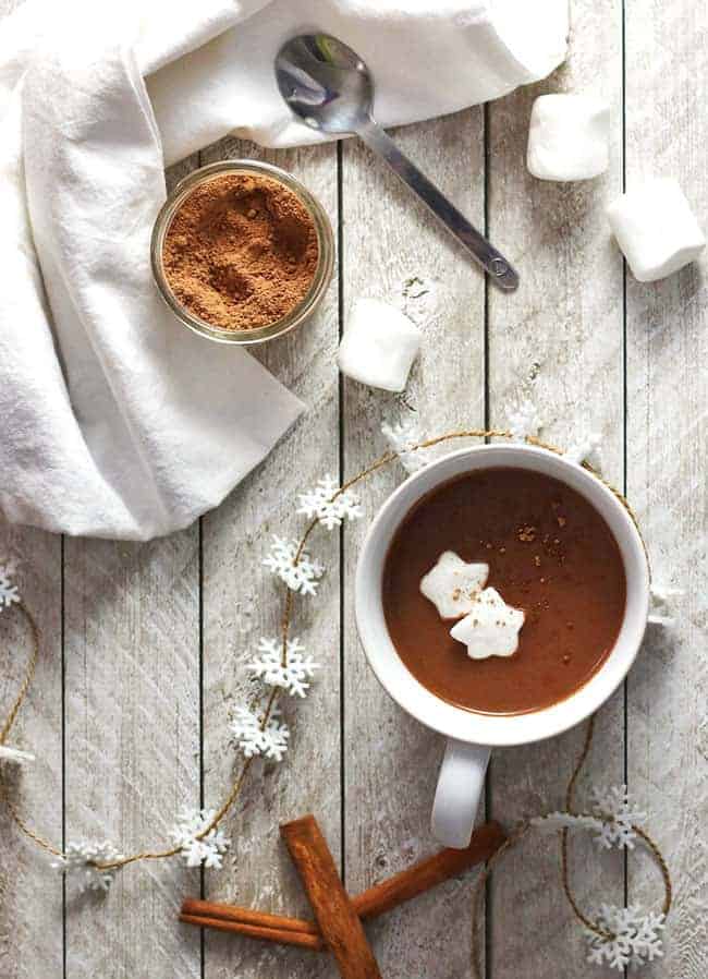 overhead shot of a white mug of vegan Gingerbread Hot Chocolate on a distressed wooden table with a snowflake garland and whole cinnamon sticks.