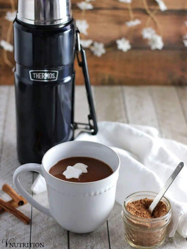 white mug of vegan Gingerbread Hot Chocolate with a large thermos in the background and a small mason jar of cacao powder in the foreground.