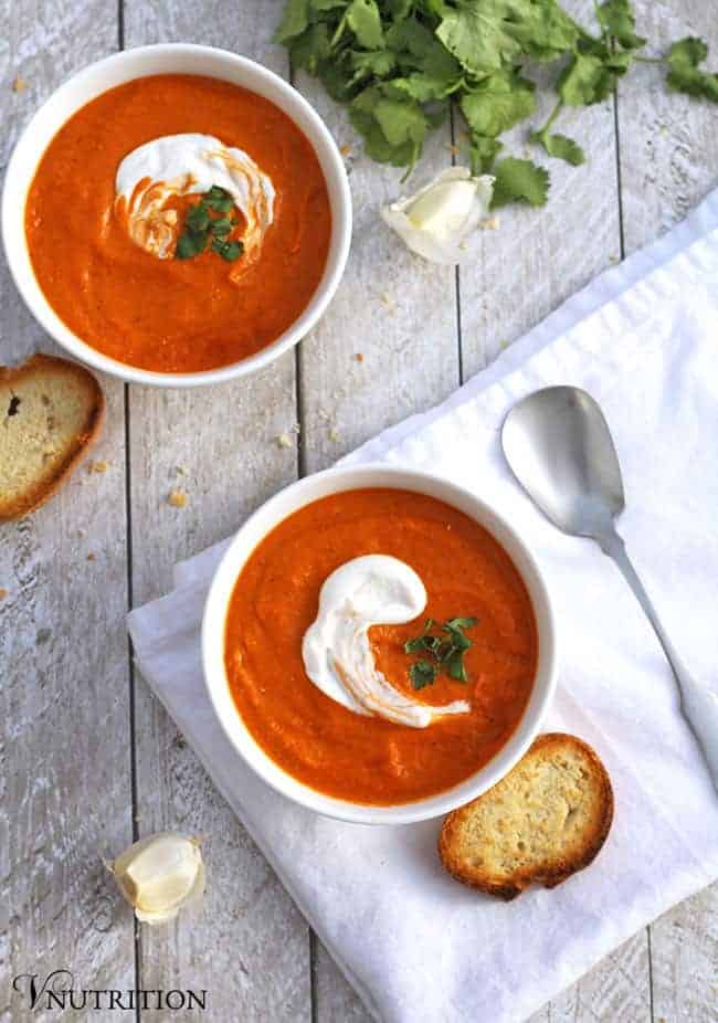 overhead shot of 2 bowls of vegan chickpea tomato soup with spoon, garlic cloves and bread on a table