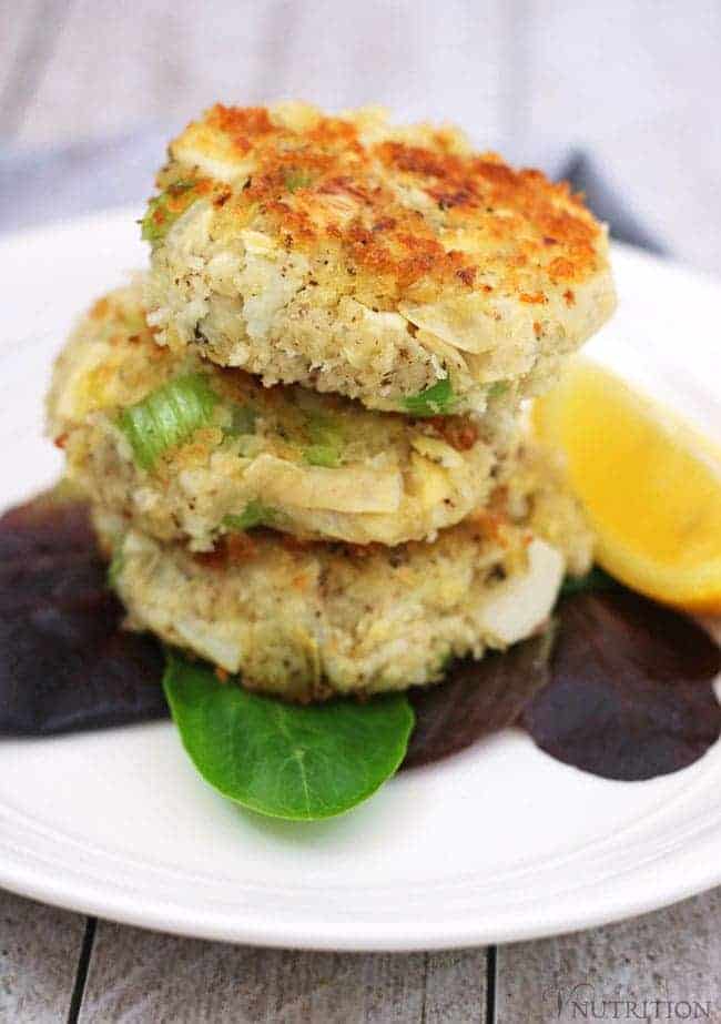 3 Vegan Crabless crab Cakes in a stack on a round white plate with lemon wedges.