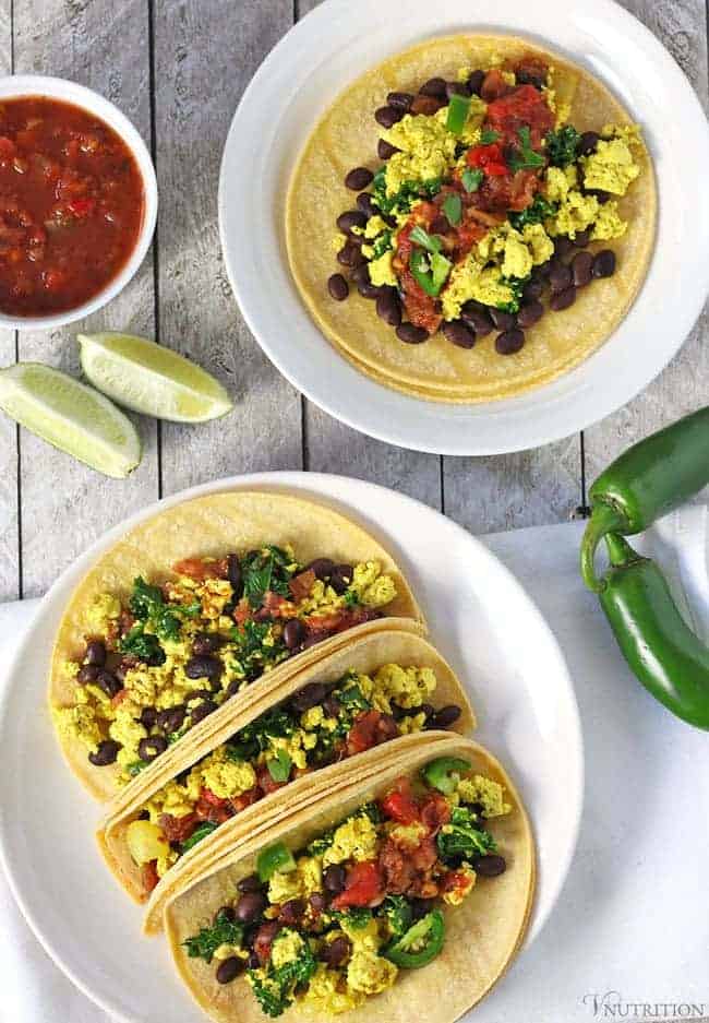 overhead shot of two plates of vegan breakfast tacos topped with salsa with a bowl of salsa, some lime quarters, and jalapeños for garnishing. 