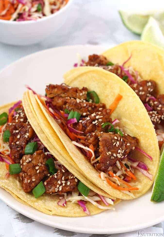 3 tempeh tacos on corn tortillas on a white plate.