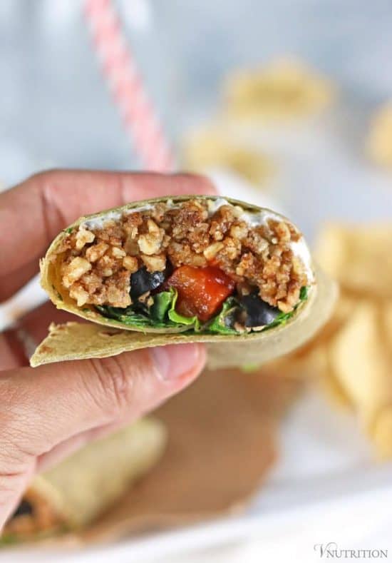 hand holding Vegan burrito filled with walnut taco meat, with chips and water glass with straw in background.