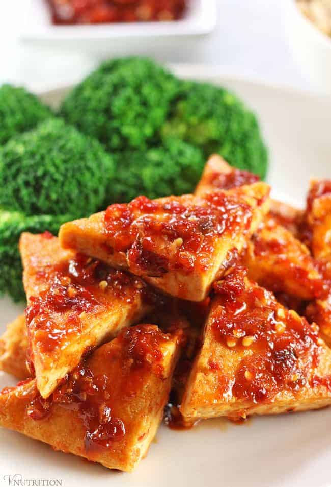 closeup of tofu in chili garlic sauce with broccoli and chili sauce in background