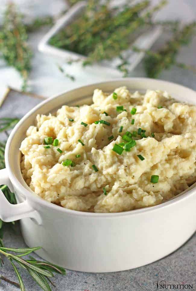 Vegan Cauliflower Mashed Potatoes in white serving bowl topped with chives on grey napkin with fresh rosemary and thyme in the background
