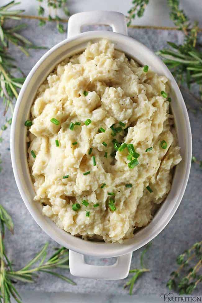 overhead shot of Vegan Cauliflower Mashed Potatoes in white serving bowl with fresh herbs surrounding the bowl