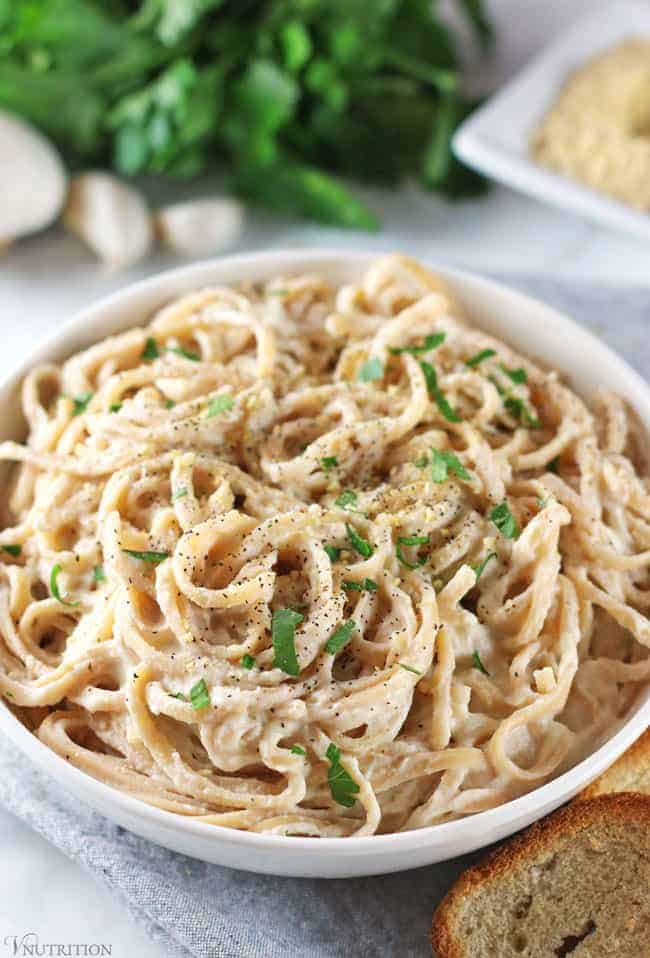 close up of vegan cauliflower alfredo sauce topped with pepper and parsley with more parsley, garlic and vegan Parmesan in background