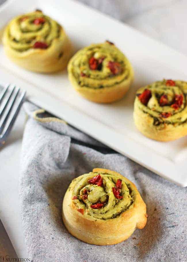 sun dried tomato and pesto Pinwheel appetizer on a grey napkin in foreground and a plate of three Christmas Pinwheels in background with tongs of a fork on the side 