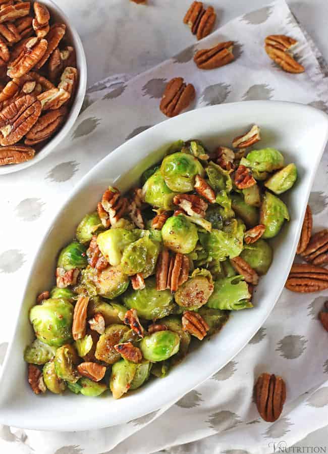 overhead shot of maple mustard brussels sprouts in an oblong serving dish on a white tablecloth scattered with pecans.