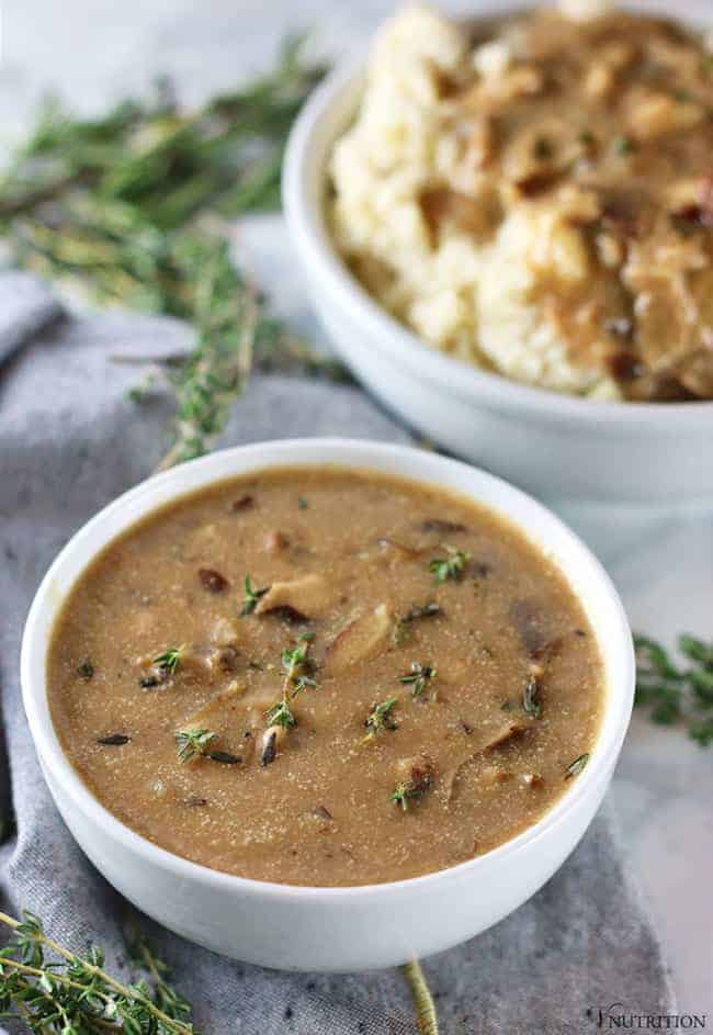 bowl of shiitake mushroom gravy with herbs around bowl and bowl of mashed potatoes with gravy in background