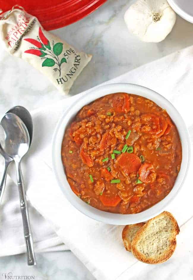 Overhead shot of vegan lentil soup in a white bowl on a table with two silver spoons and some toast.