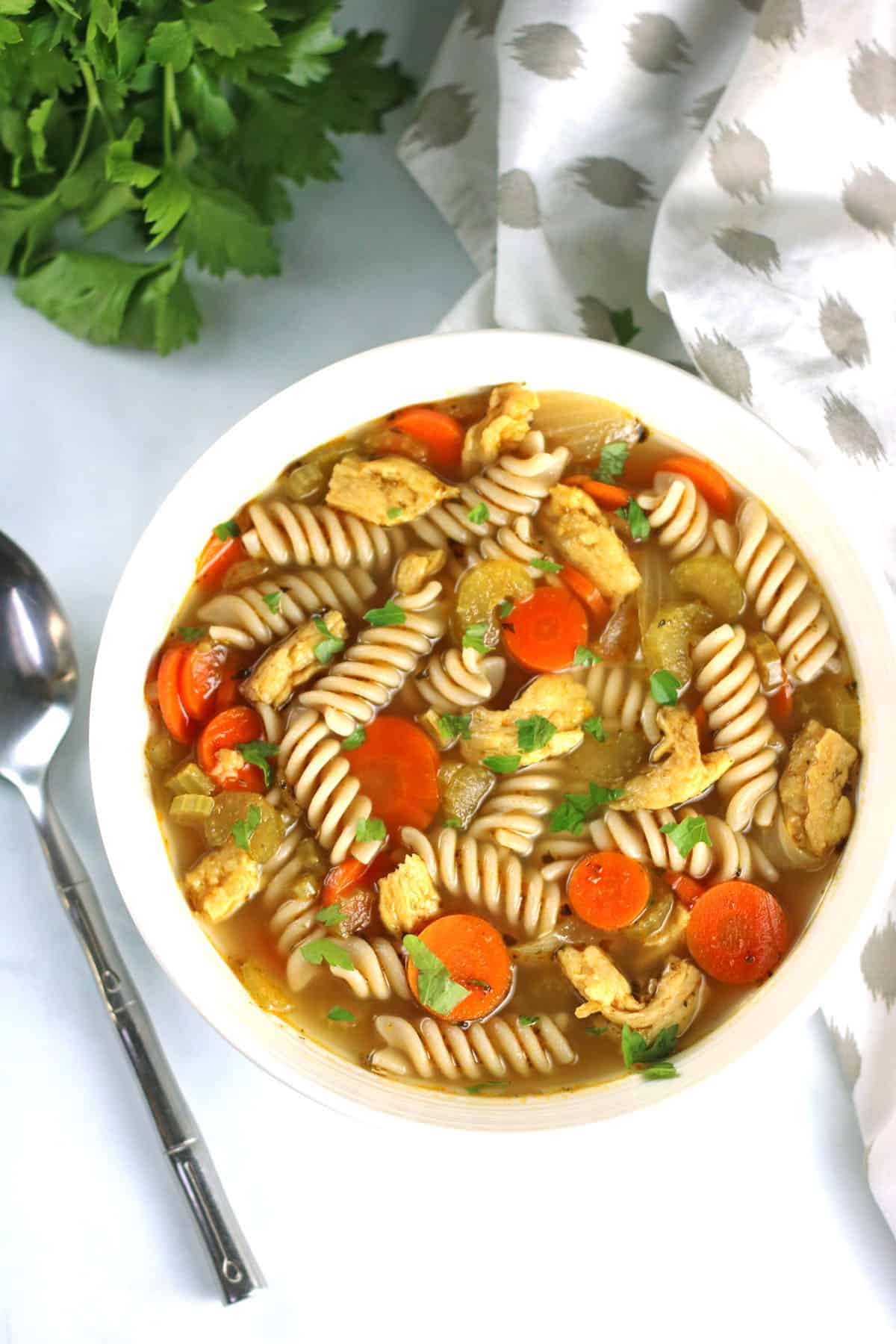 overhead shot of vegan chicken noodle soup in a white bowl topped with fresh parsley.