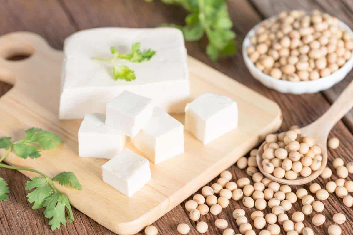 block of tofu on a wooden cutting board with a few cubes that have been cut, some cilantro leaves as garnish, and a scattering of soybeans