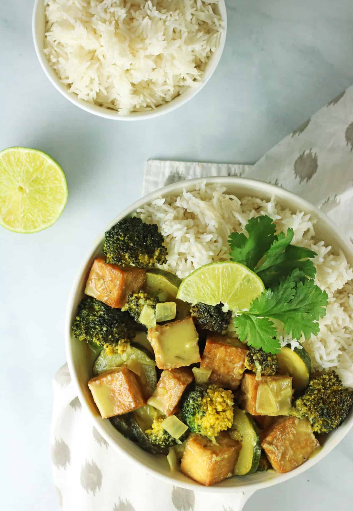 overhead shot of vegan Green Curry Tofu in a white bowl on a white table with a bowl of white rice.