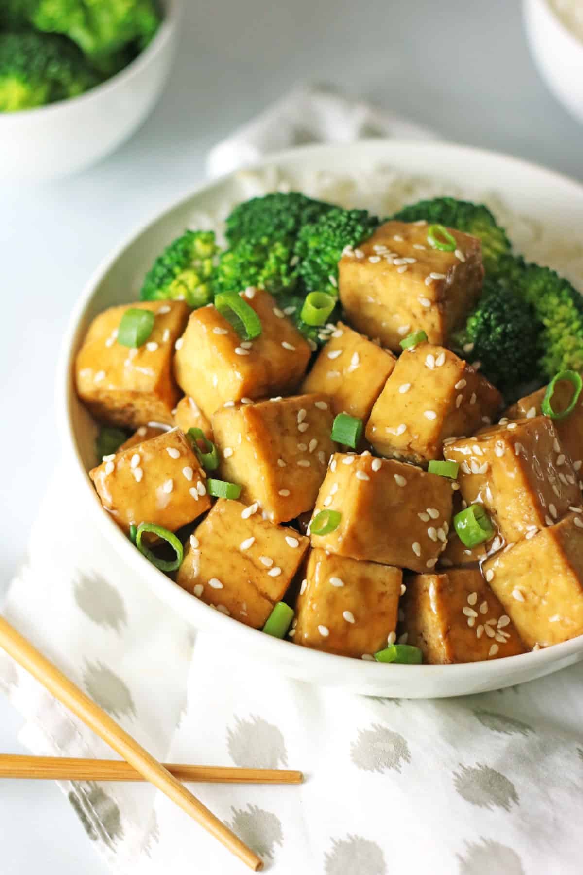 45 degree angle shot of white bowl with crispy baked vegan teriyaki tofu cubes, steamed broccoli and white rice garnished with sesame seeds and scallions.