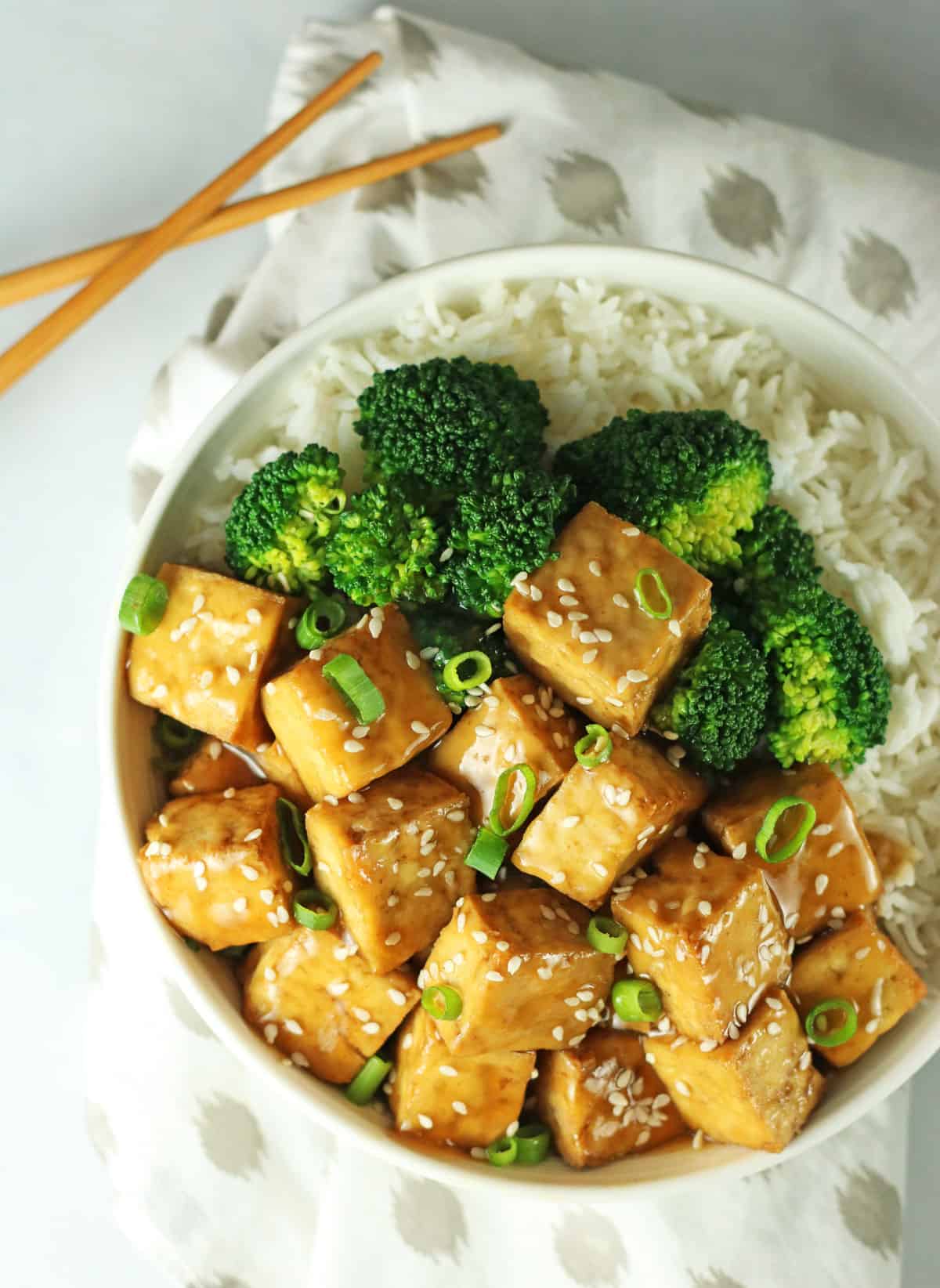 close up shot of a white bowl filled with sticky rice, steamed broccoli, and crispy baked vegan teriyaki tofu cubes.