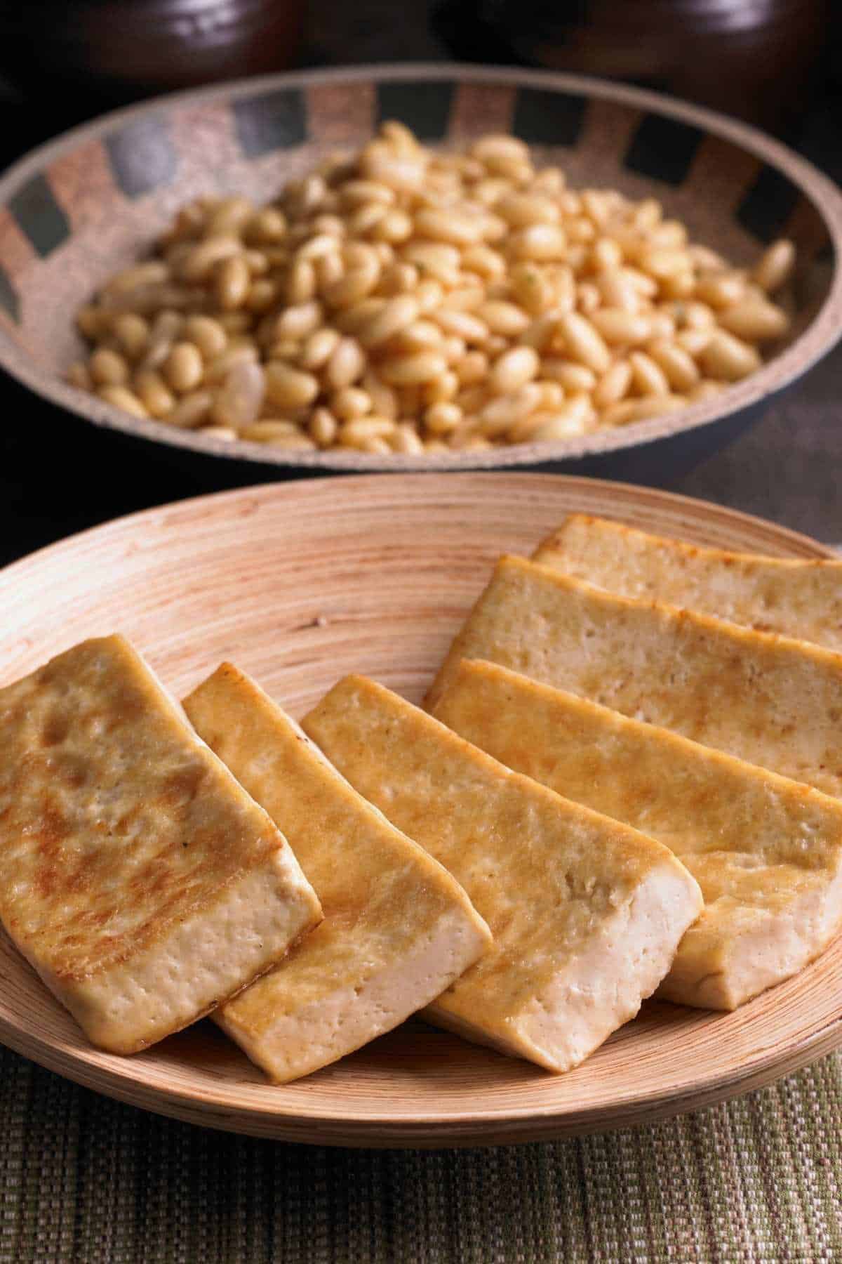 cooked slices of tofu on a plate with soybeans in a bowl in the background