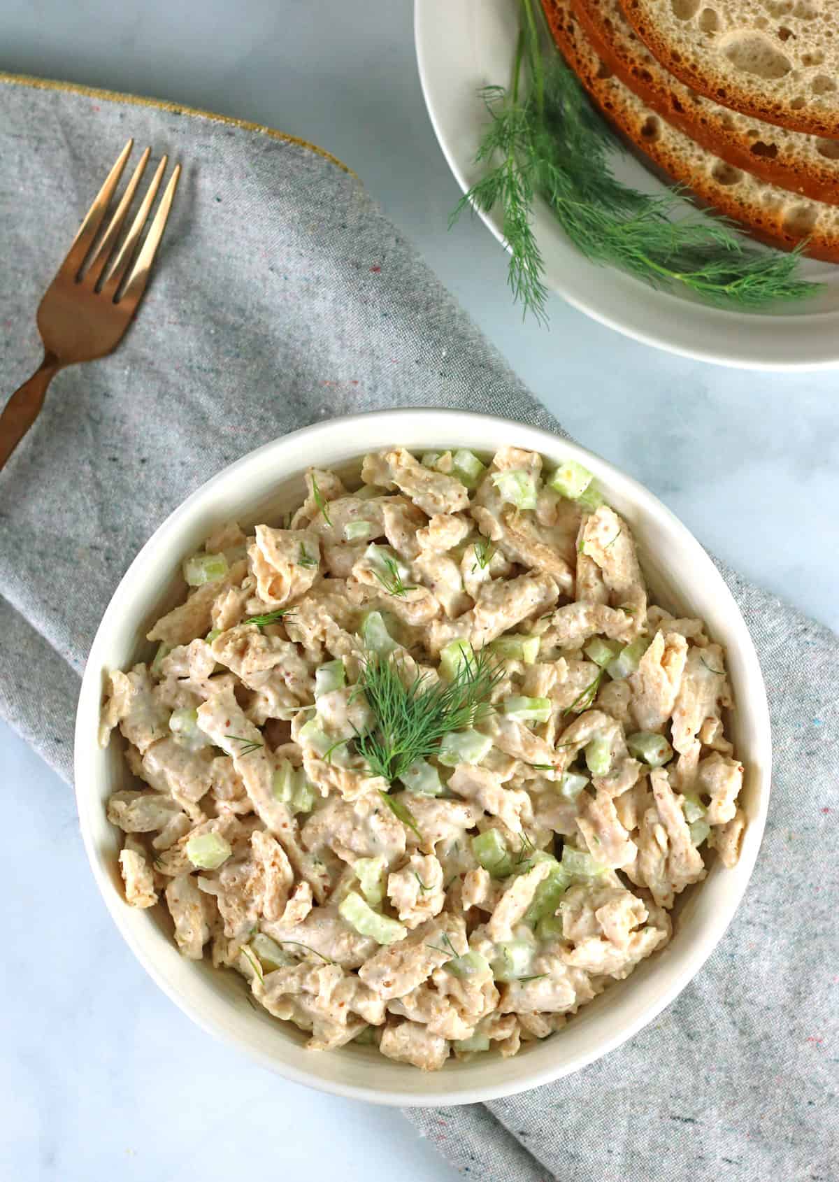 overhead of vegan chicken salad in white bowl with gray napkin, gold fork, and bread.