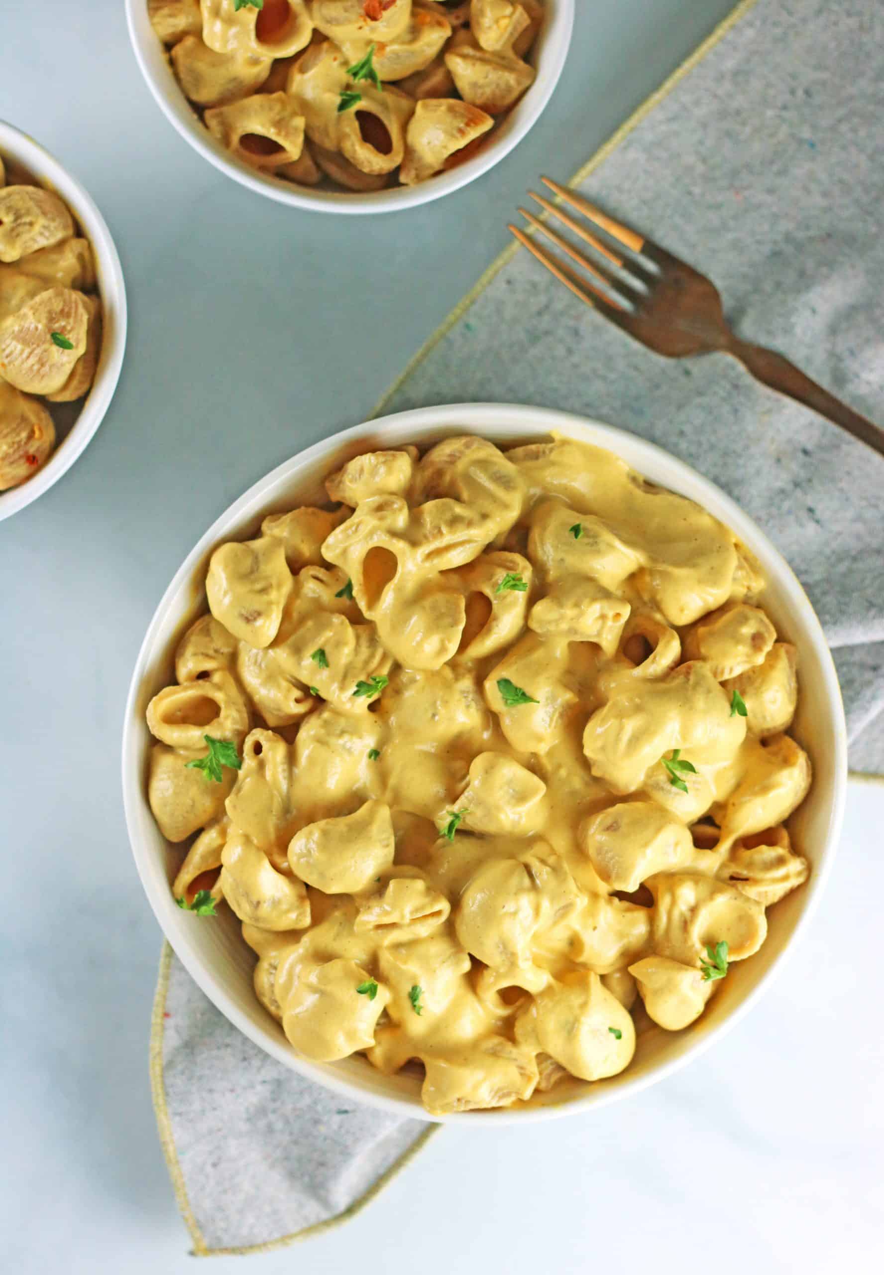 large bowl of vegan butternut squash mac and cheese with gold fork, grey napkin and 2 smaller bowls of pasta.