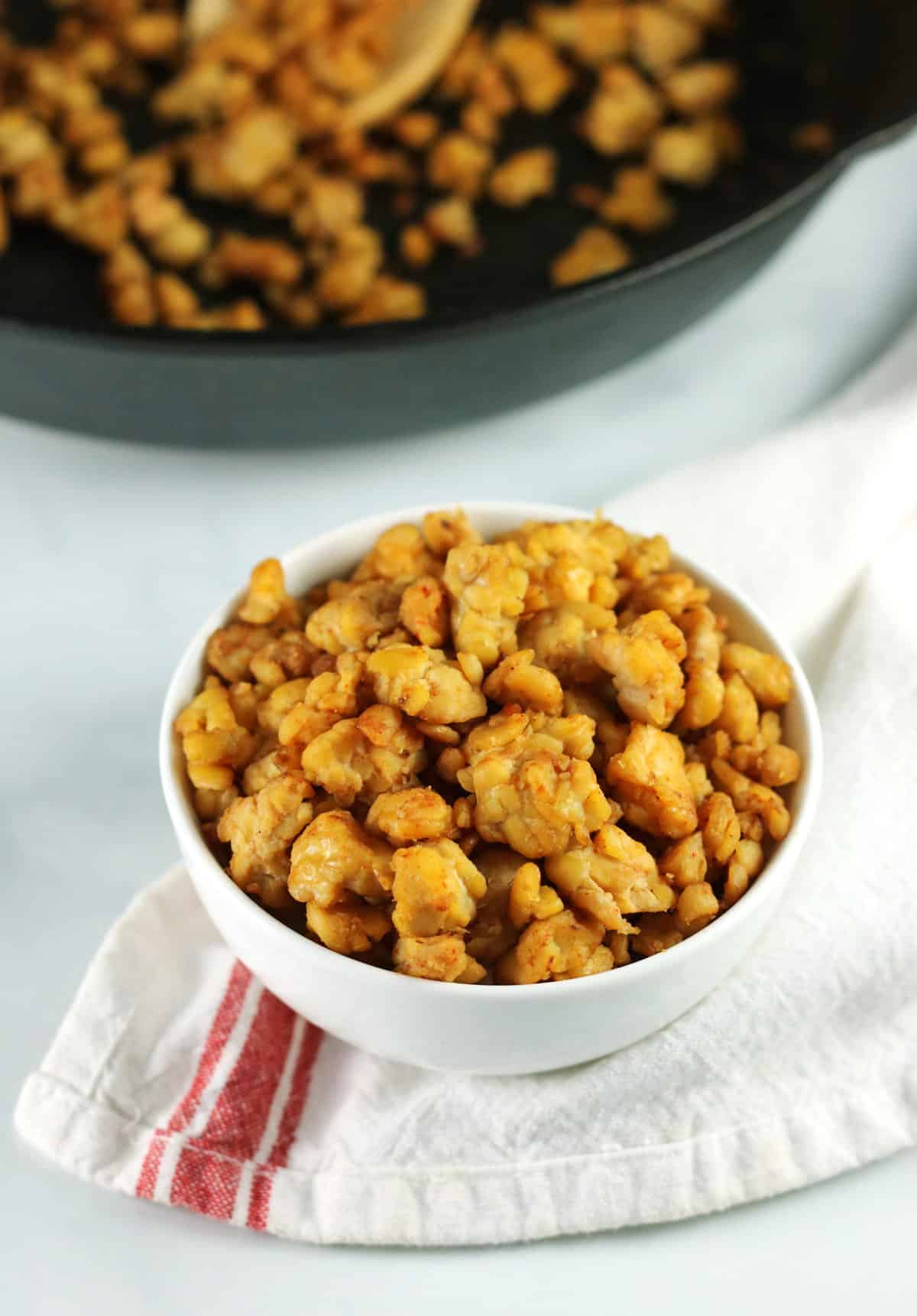 tempeh in white bowl with white and red napkin and skillet in background