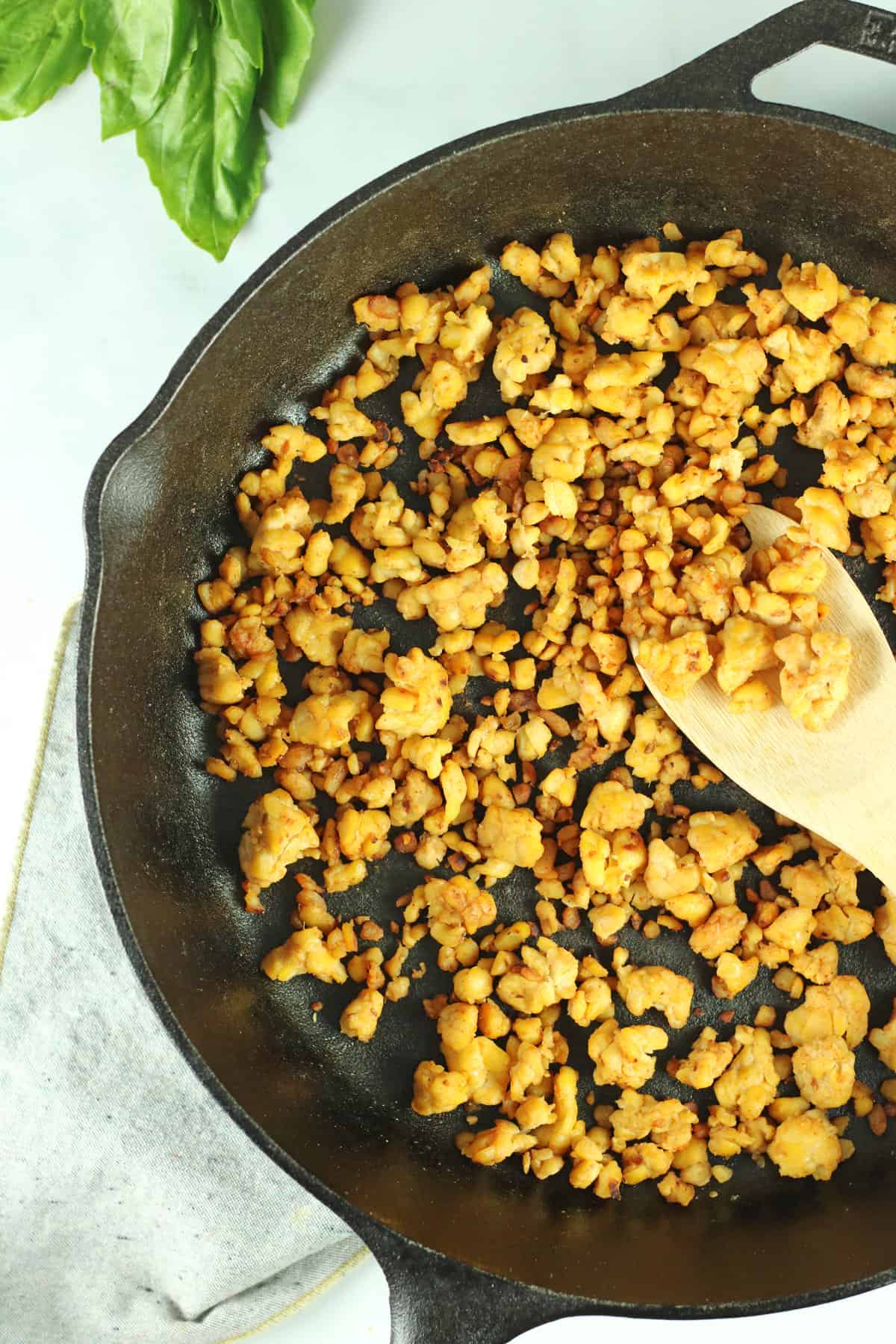 tempeh-based meatless crumbles in cast iron skillet with wooden spook and basil on side.