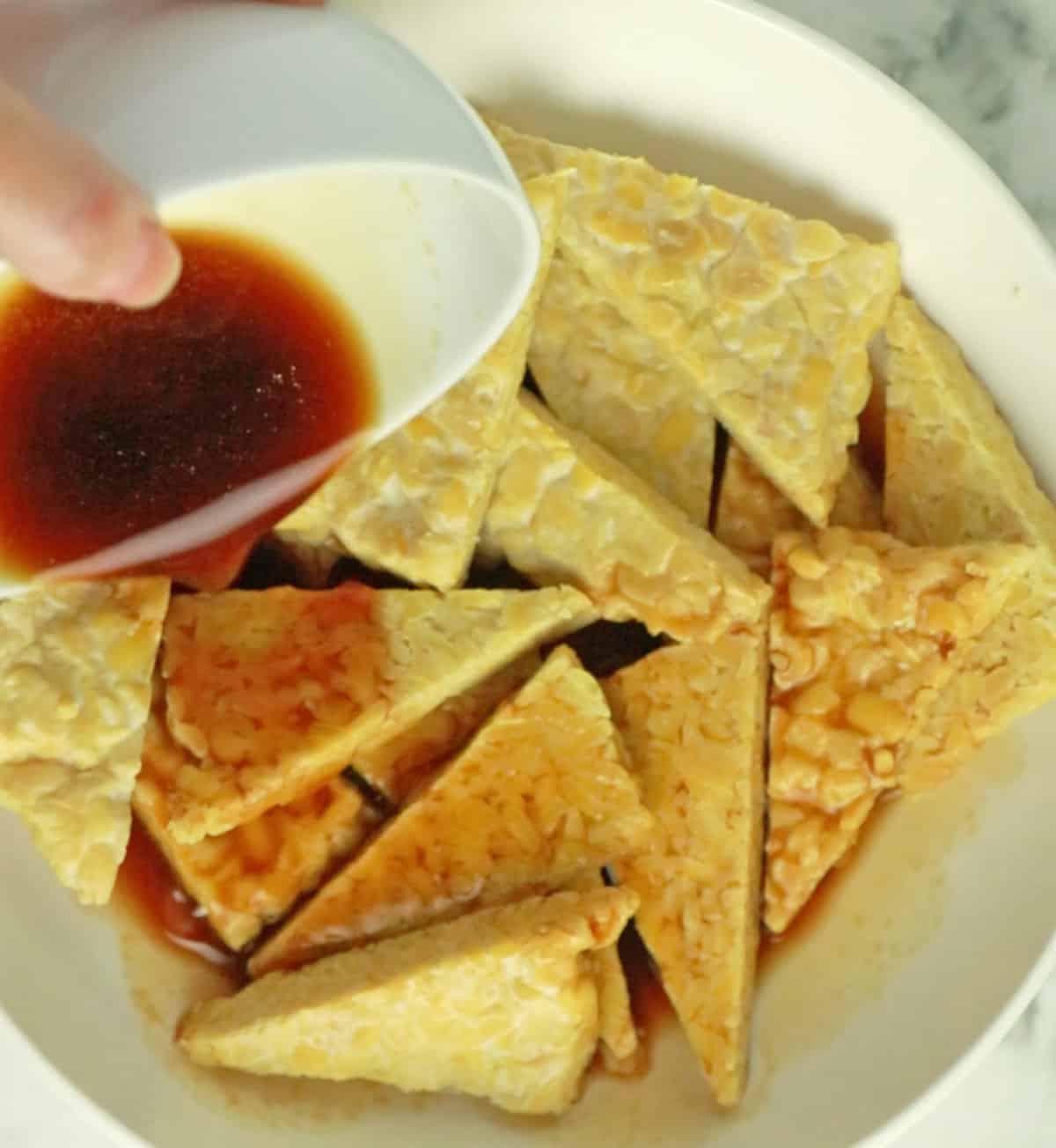 pouring marinade over tempeh in white bowl.