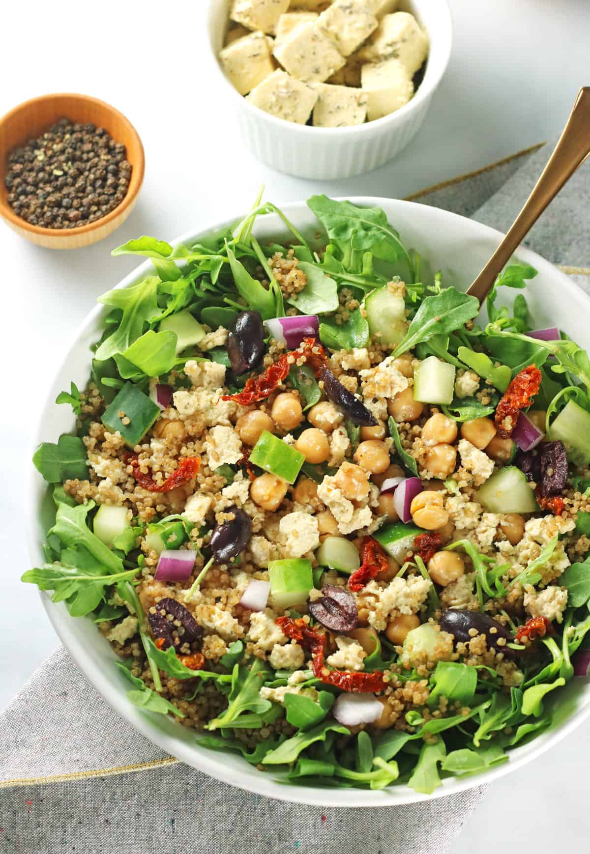 mixed greek quinoa salad in white bowl with gold fork, black peppercorns, and tofu feta