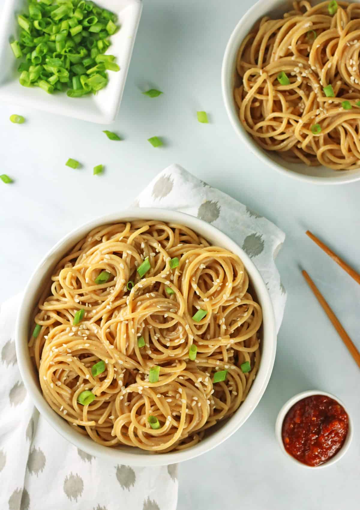 overhead shot of two bowls of spicy sesame noodles  with small bowl of scallion and chopsticks.